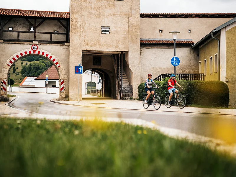 Zwei Radfahrer fahren an einem historischen Stadttor mit rotem Dach und Durchfahrt vorbei.