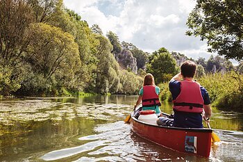 Paddler bei den Zwölf Aposteln Eine Frau und ein Mann paddeln auf der Altmühl in einem roten Kanu mit dem Rücken zu uns. Das Ufer ist von grünen Bäumen gesäumt.