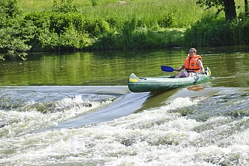 Ein Bootsfahrer mit oranger Rettungweste und Paddel steht mit seinem grünen Kanu am Anfang der Bootstreppe in der Altmühl. Das Wasser rauscht hier an dem steinernen Wehr hinunter. Die Bootsrutsche ist eine Vertiefung für die Fische und auch Boote. Das Ufer ist sattgrün auf dem die Sonne scheint.