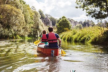 Bootwanderer auf der Altmühl Ein Mann und eine Frau mit dem Rücken zu uns, sitzen in einem roten Kanu auf der Altmühl. Die gelben Paddel erzeugen leichte Wellen auf der Wasseroberfläche. im Hintergrund ist das grüne Ufer mit prächtigen Bäumen zu sehen und rechts blitzt der blaue Himmel durch.