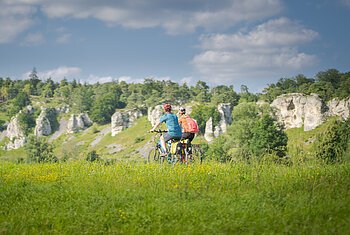 Zwei Personen fahren mit Fahrrädern auf einem grasbewachsenen Feld vor einer Felslandschaft.