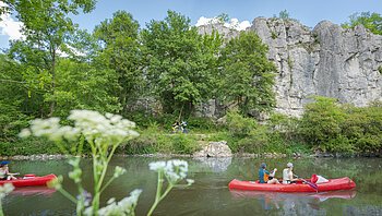 Zwei rote Kanus auf einem Fluss, im Hintergrund Felsen und Bäume, im Vordergrund unscharfe Blumen.