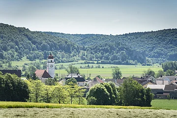 Landschaft mit Dorf, Kirchturm und bewaldeten Hügeln im Hintergrund.