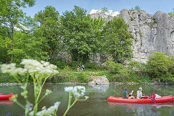 Zwei rote Kanus auf einem Fluss, im Hintergrund Felsen und Bäume, im Vordergrund unscharfe Blumen.
