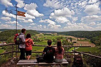 Vier Personen mit Rucksäcken blicken von einem Aussichtspunkt auf grüne Felder und bewaldete Hügel unter blauem Himmel.
