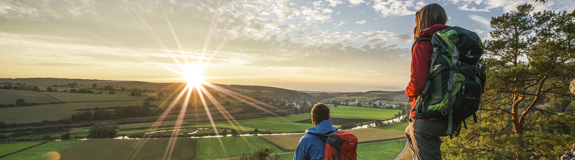 Zwei Wanderer mit Rucksäcken auf Felsen, Blick auf grüne Felder und Sonnenuntergang am Horizont.