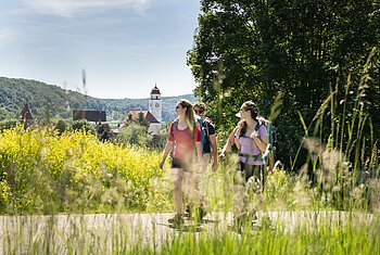 Drei Wanderer mit Rucksäcken auf Weg vor gelben Blumen und Dorf mit Kirchturm im Hintergrund