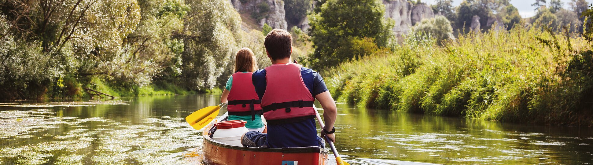 Zwei Personen mit roten Schwimmwesten paddeln in einem roten Kanu auf einem Fluss, umgeben von grüner Natur.