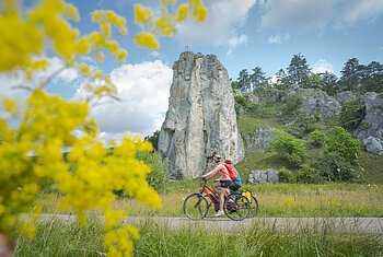 Zwei Radfahrer fahren auf einem Weg vor einem hohen Felsen mit Kreuz, gelbe Blumen im Vordergrund, bewölkter Himmel