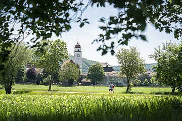 Zwei Personen gehen auf einem Weg durch eine grüne Wiese, im Hintergrund ein Kirchturm und Bäume.
