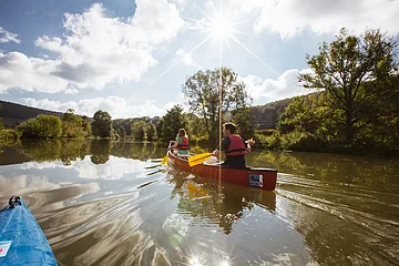 Zwei Personen paddeln in einem roten Kanu auf einem ruhigen Fluss bei sonnigem Himmel mit Wolken.