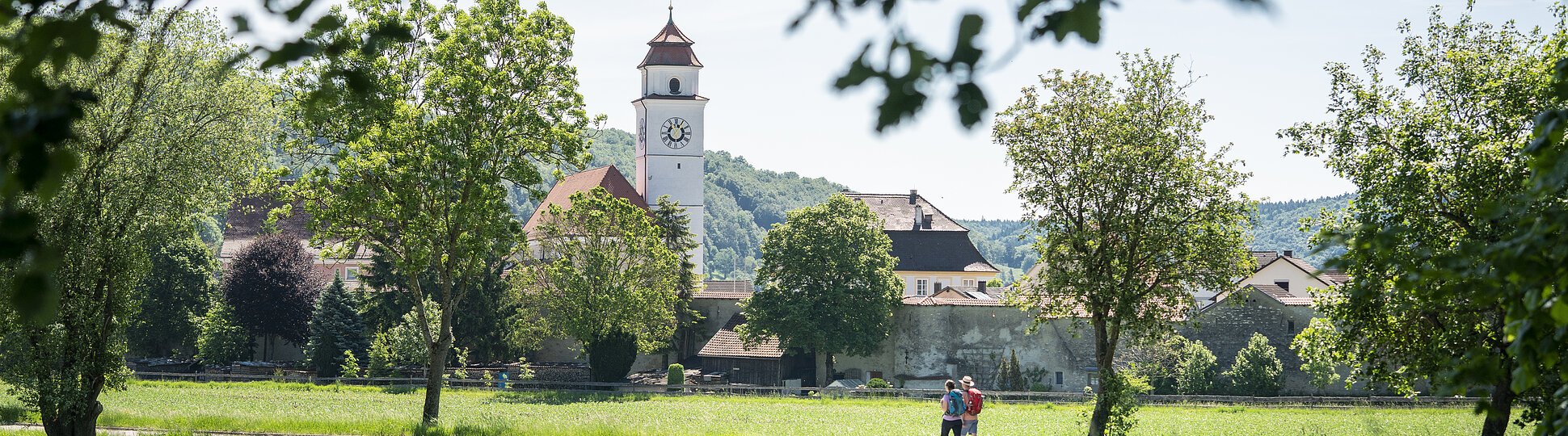 Zwei Personen gehen auf einem Weg durch eine grüne Wiese, im Hintergrund ein Kirchturm und Bäume.