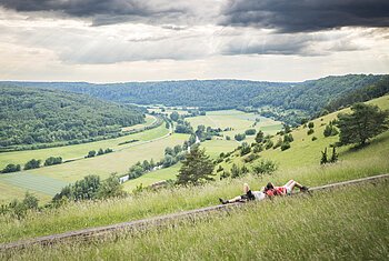 Zwei Wanderinnen liegen auf einer langen Holzbank und schauen ins weitläufige grüne Altmühltal.