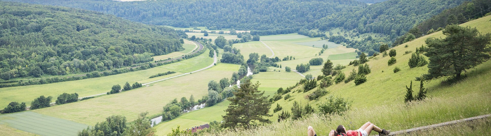 Zwei Wanderinnen liegen auf einer langen Holzbank und schauen ins weitläufige grüne Altmühltal.