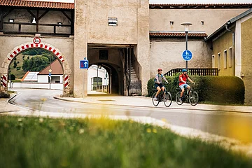 Zwei Radfahrer fahren an einem historischen Stadttor mit rotem Dach und Durchfahrt vorbei.