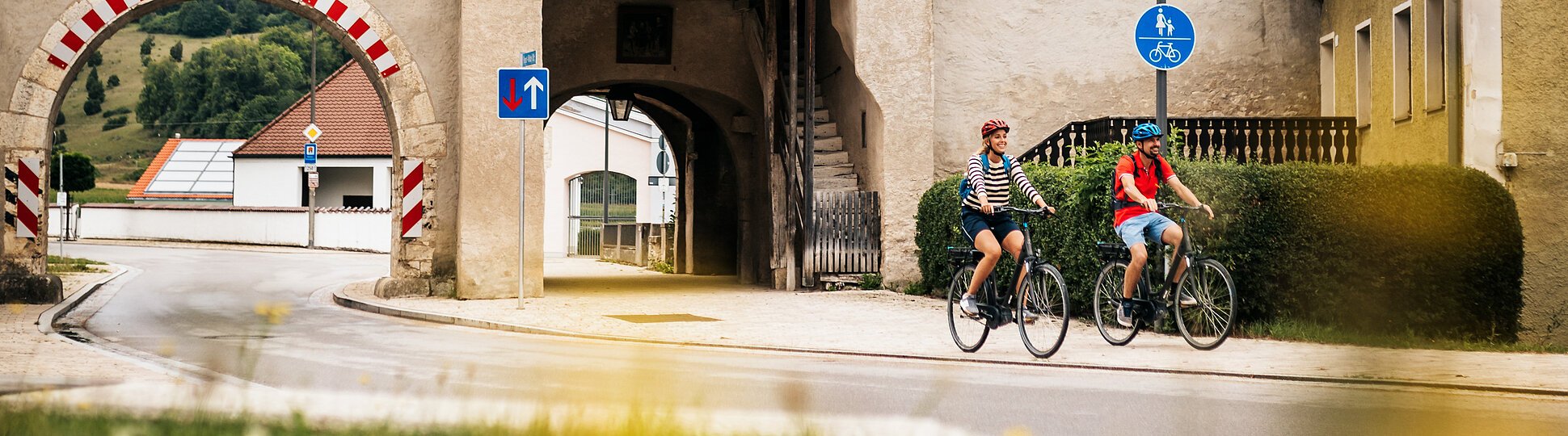 Zwei Radfahrer fahren an einem historischen Stadttor mit rotem Dach und Durchfahrt vorbei.