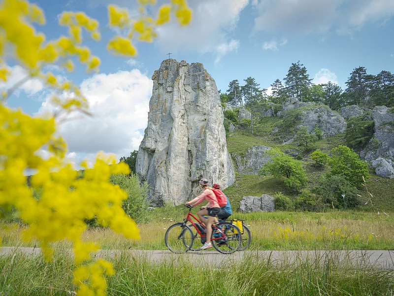 Radfahrer am Burgsteinfelsen Zwei Radfahrer fahren auf einem Weg vor einem hohen Felsen mit Kreuz, gelbe Blumen im Vordergrund, bewölkter Himmel
