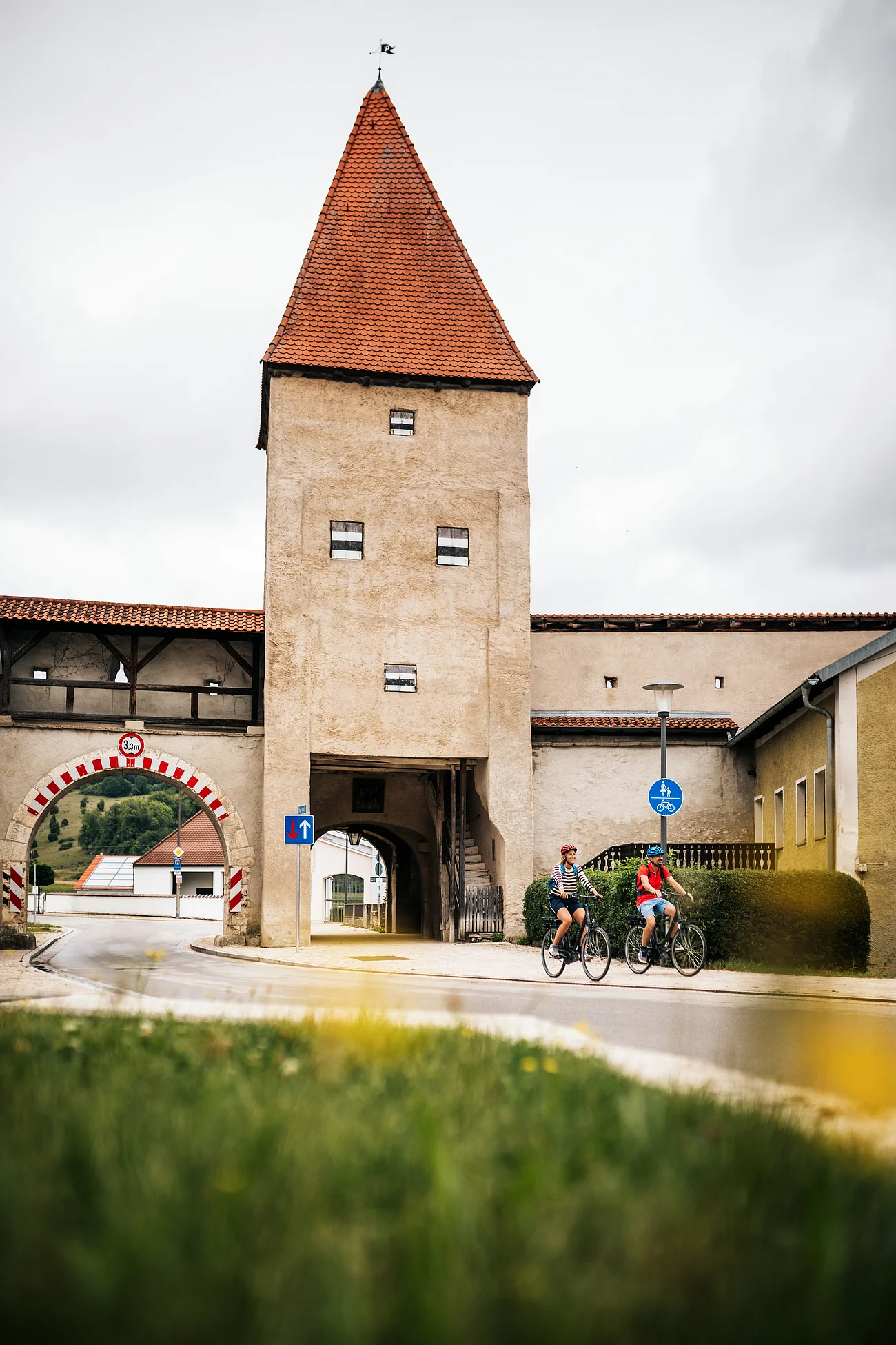 Altmühltal-Radweg (Dollnstein) Zwei Radfahrer fahren an einem historischen Stadttor mit rotem Dach und Durchfahrt vorbei.