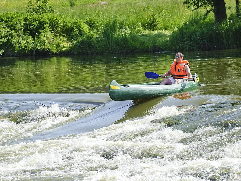 Ein Bootsfahrer mit oranger Rettungweste und Paddel steht mit seinem grünen Kanu am Anfang der Bootstreppe in der Altmühl. Das Wasser rauscht hier an dem steinernen Wehr hinunter. Die Bootsrutsche ist eine Vertiefung für die Fische und auch Boote. Das Ufer ist sattgrün auf dem die Sonne scheint.