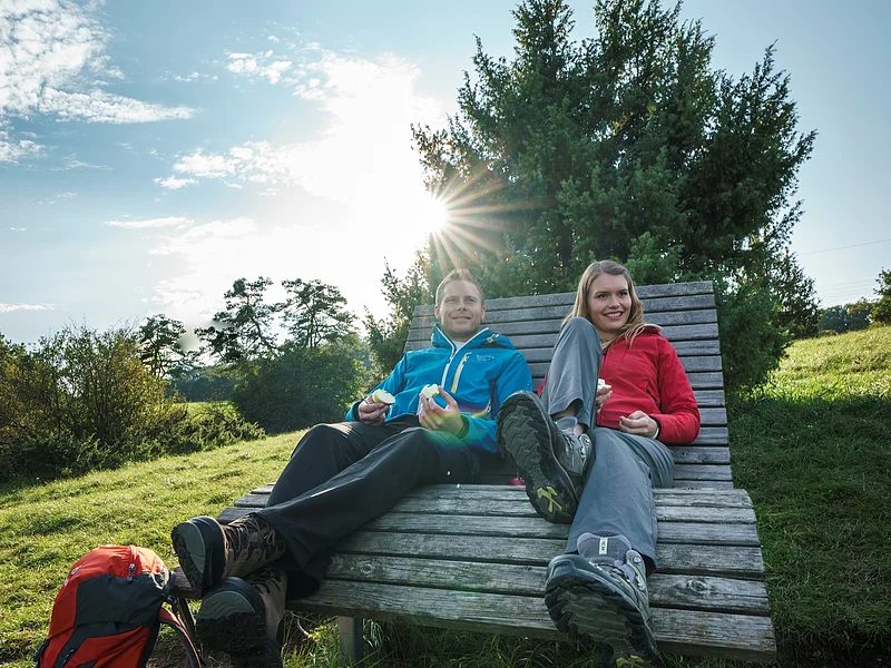 Wanderer auf Liegebank bei Hagenacker Ein Wanderpärchen sitzt auf einer Liegebank aus Holz. Der Mann mit blauer Jacke hält die Brotzeit in der Hand. Neben ihm sitzt eine blonde Frau mit roter Jacke. Hinter ihnen ist ein großer Busch und die Sonne scheint beiden in den Rücken.