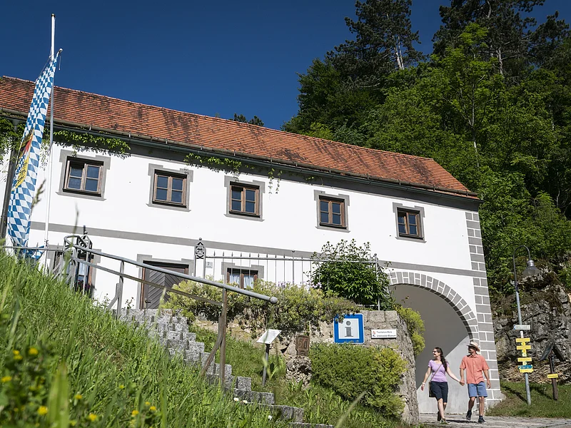Weißes Gebäude mit rotem Ziegeldach, bayerischer Flagge und zwei Personen vor einem Torbogen bei Sonnenschein