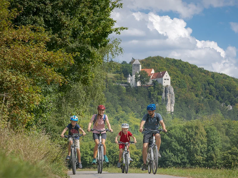 Familie mit zwei Kindern fährt auf Fahrrädern auf einem Weg vor bewaldetem Hügel mit Burg im Hintergrund.