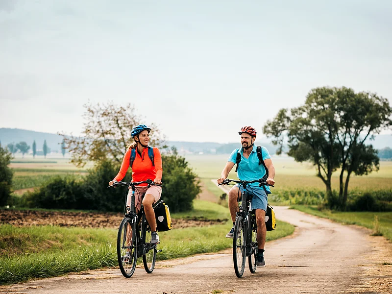 Eine Frau und ein Mann radeln auf dem Altmühltal-Radweg bei Dittenheim. Beide tragen einen Fahrradhelm und einen Rucksack. Der Radweg wird von Gras und grünen Bäumen gesäumt.