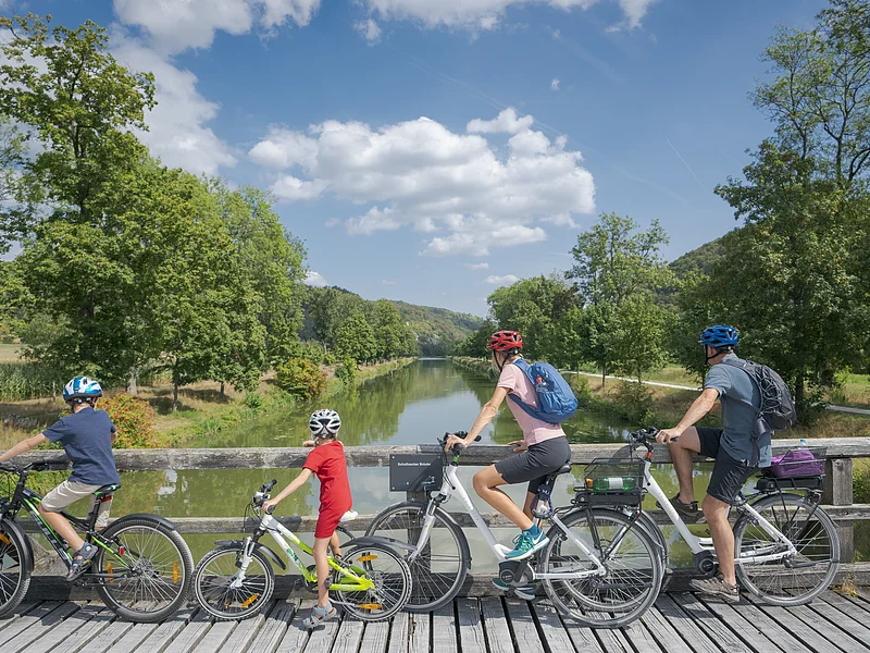 Vier Personen mit Fahrrädern auf einer Holzbrücke mit Blick auf einen Fluss und grüne Landschaft.