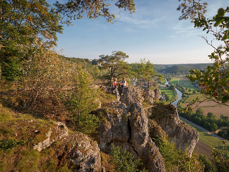 Ein Wanderpärchen, etwas entfernt, steht links auf einem Felsen an der Arnsberger Leite, die gut zu sehen ist. Dahinter erstreckt sich der Blick ins Tal. Der Himmel erstrahlt wolkenlos.
