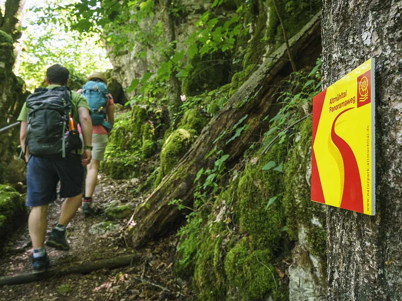 Im Vordergrund hängt ein Wanderschild des Altmühltal-Panoramaweges. Ein Wanderpärchen steigt die Treppen der Riedenburger Klamm hinauf.