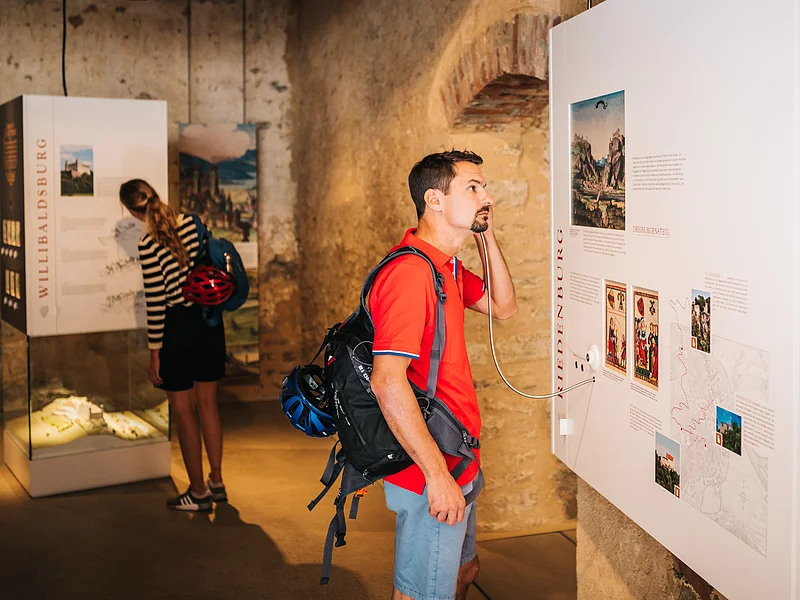 Mann mit rotem Hemd hört an Museumstafel mit Audioguide, Frau liest Ausstellungstext im Hintergrund.